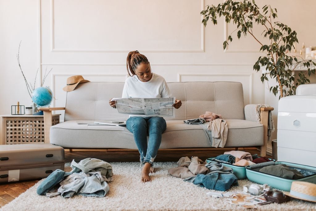 A woman sitting on the sofa, reading a newspaper, surrounded by luggage and clothes, preparing for a journey.